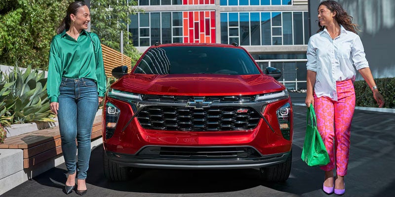 Front view of 2025 Chevy Trax and two smiling women standing in front of the car with shopping bags